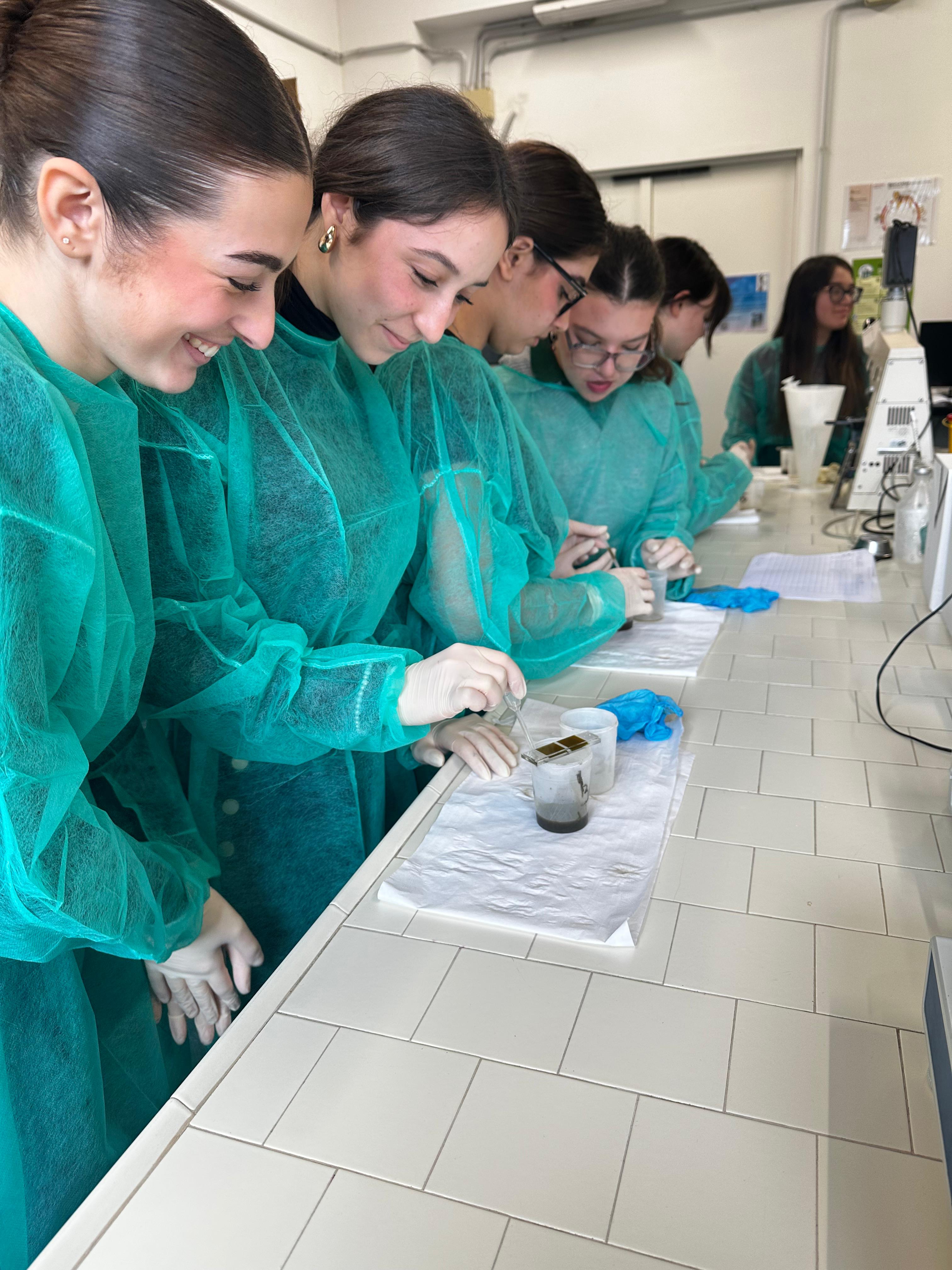 Studentesse e studenti, con camici monouso verdi e guanti, svolgono un’attività pratica di laboratorio utilizzando pipette e vetrini