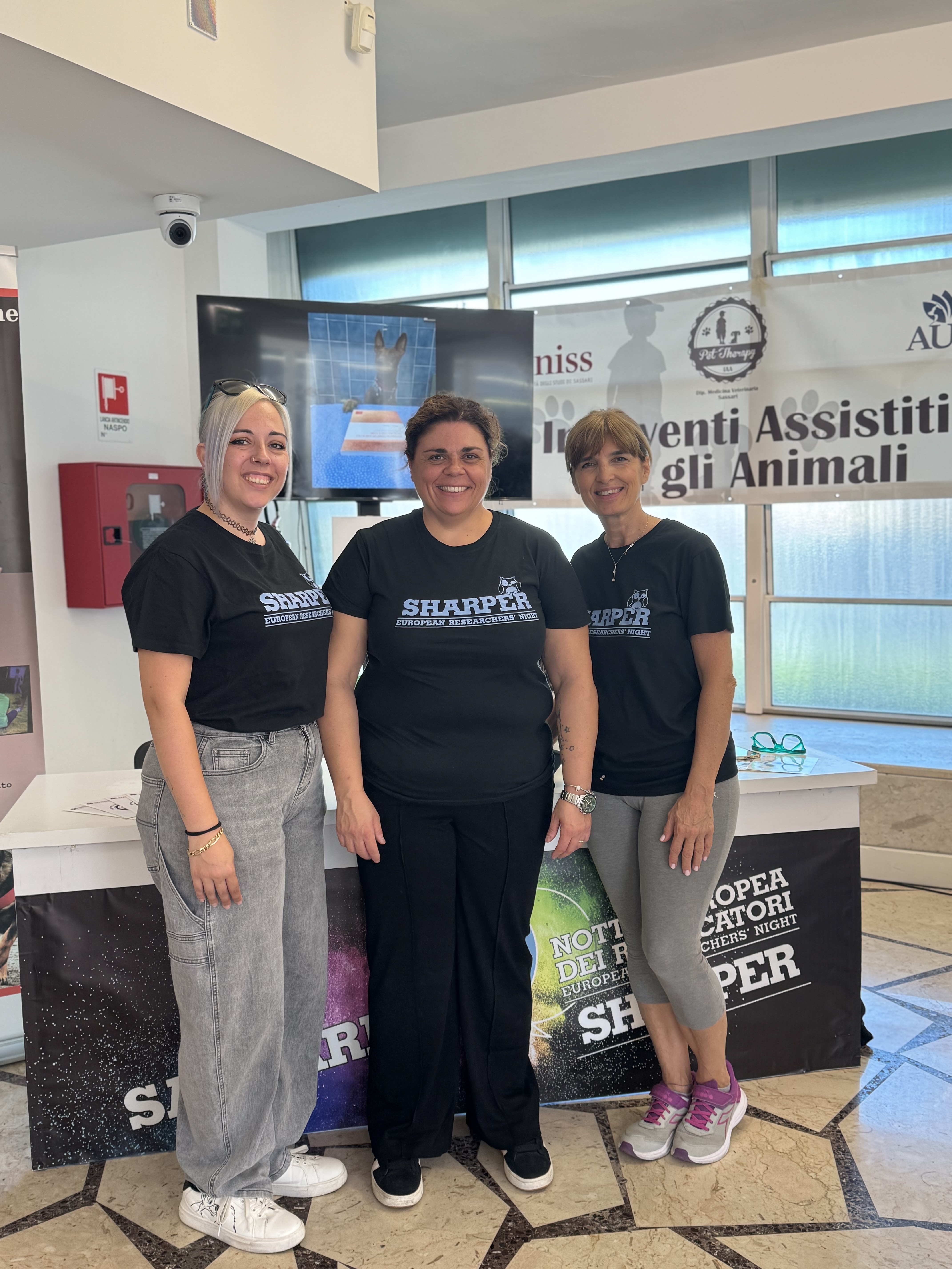 Tre membri dello staff di Vetsassari posano con la maglietta di SHARPER davanti allo stand sugli Interventi Assistiti con gli Animali.