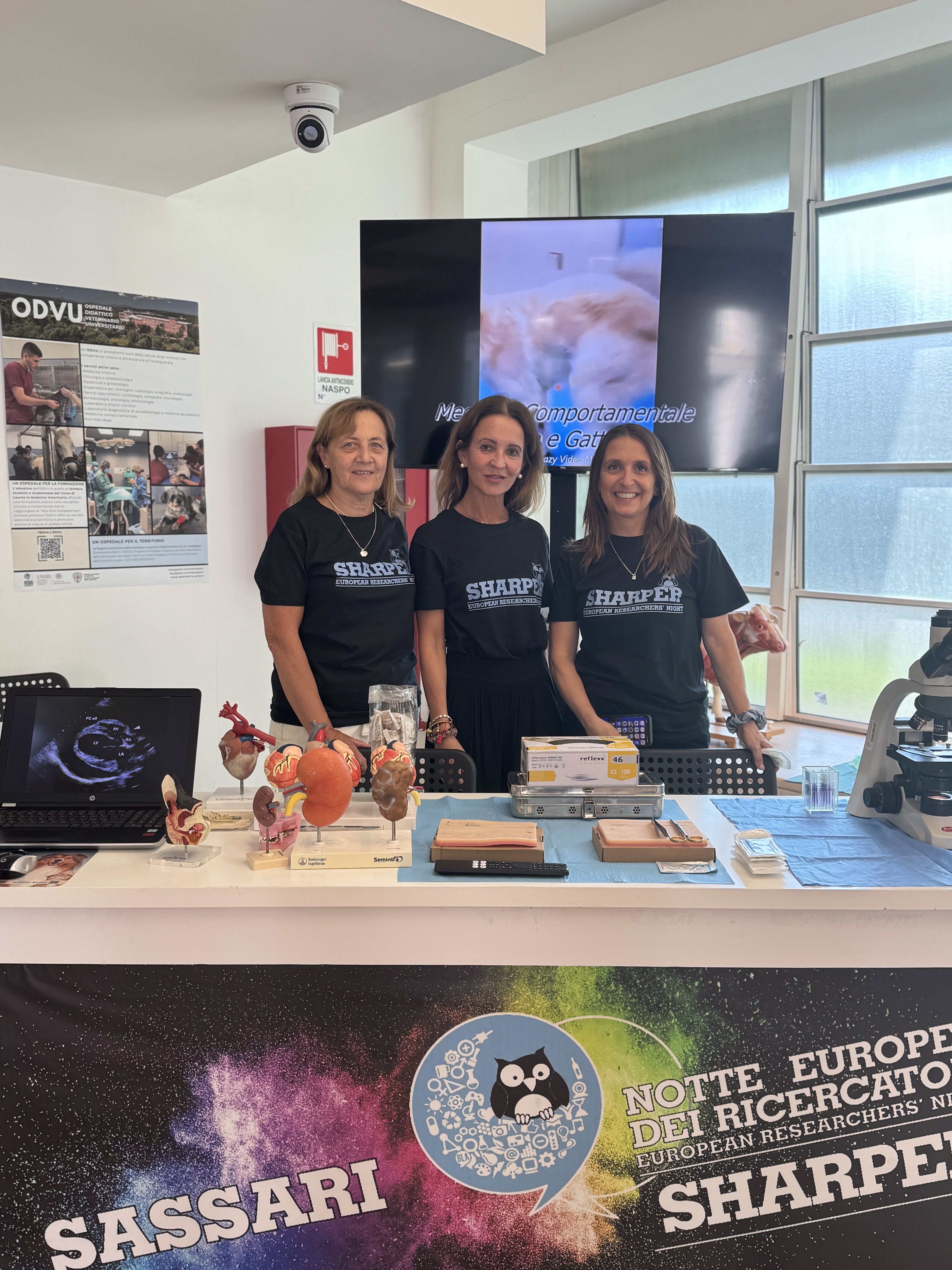 Tre membri dello staff di VetSassaru posano allo stand dell’ODVU. Sul tavolo sono esposti modelli anatomici, strumenti per esercitazioni e un microscopio.