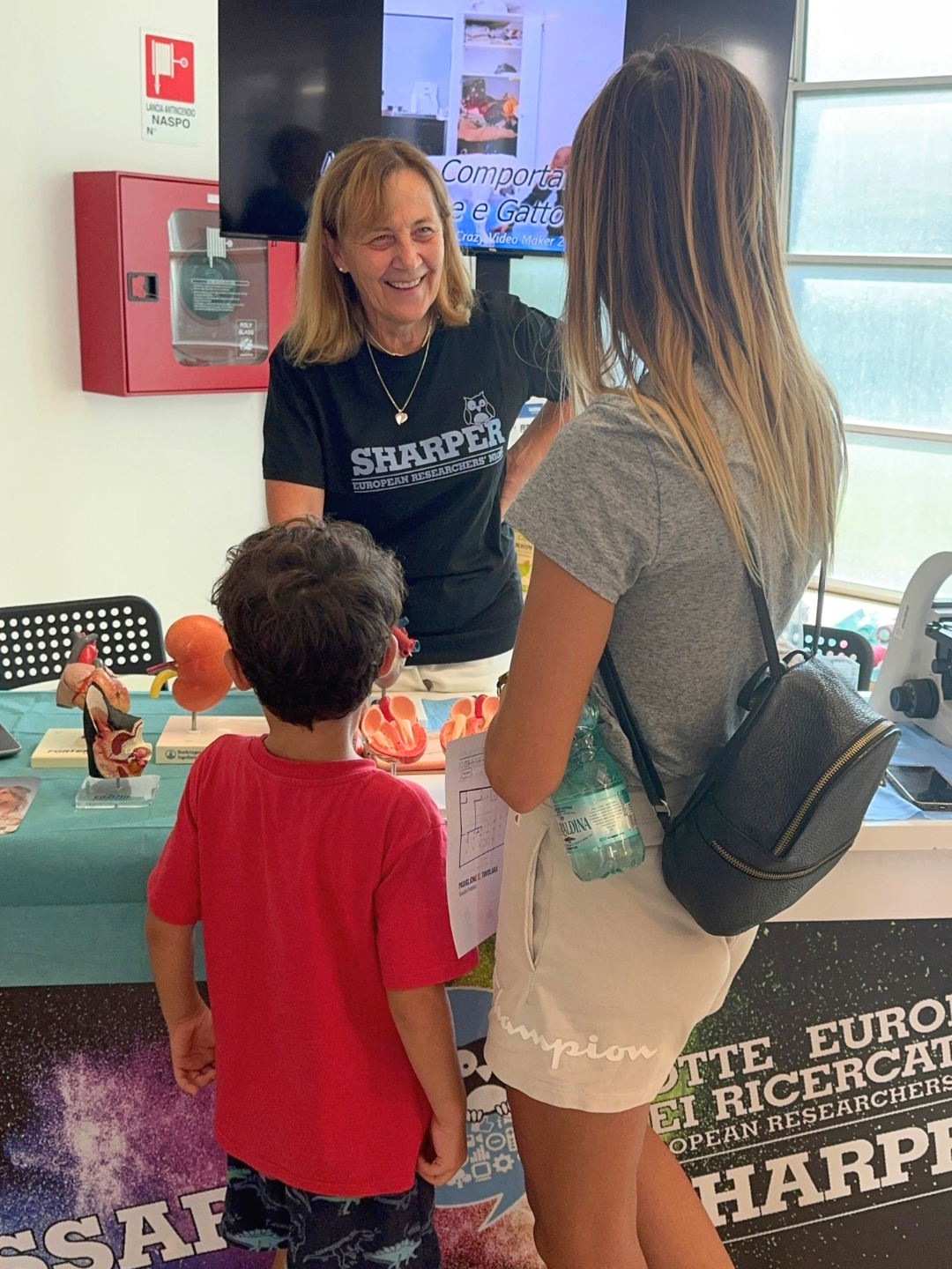conversa con una visitatrice e un bambino allo stand di VetSassari. Sul tavolo sono esposti modelli anatomici.
