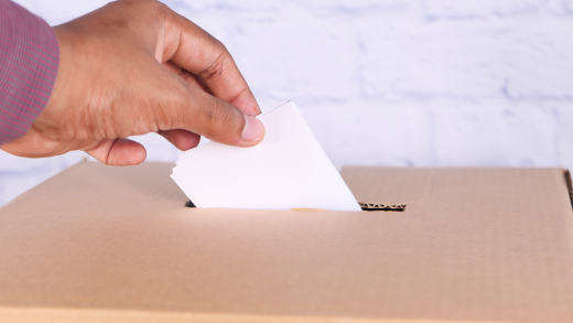 Una mano inserisce una scheda bianca in un’urna di cartone, immagine simbolica di una votazione elettorale.